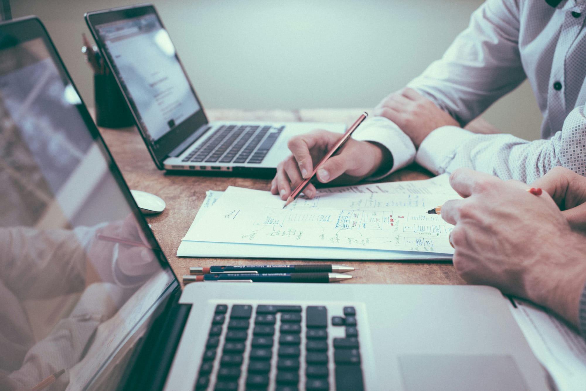 Two persons in front of laptops holding pencils over documents
